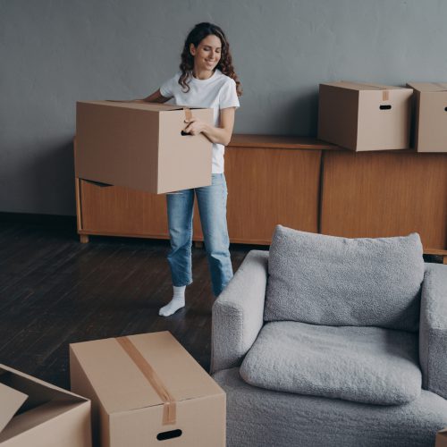 Hispanic girl is holding box and leaving apartment. Happy young woman relocates alone. Single lady moves. Real estate purchase, mortgage, delivery service ordering concept.